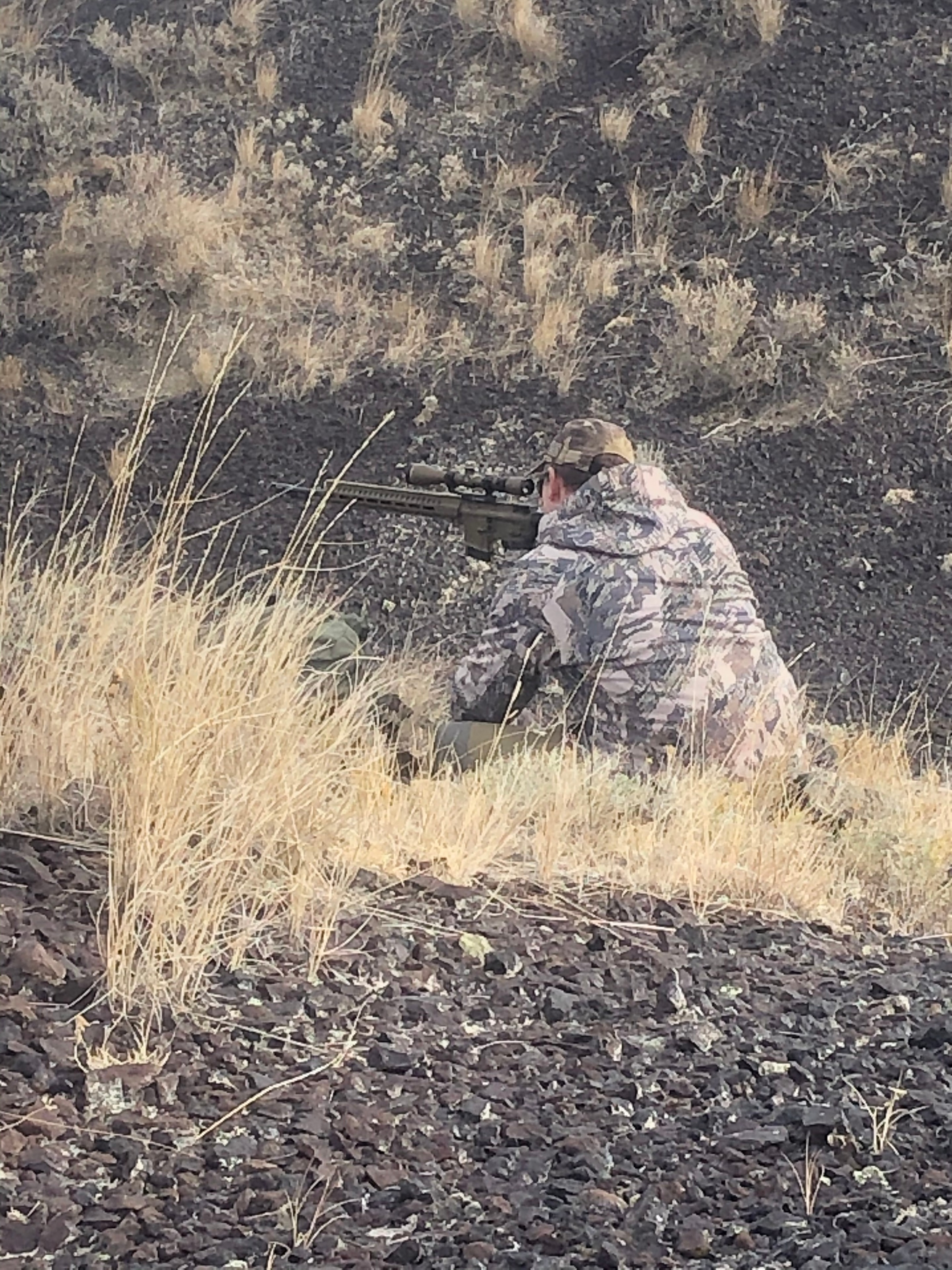Hunter in camo prone with rifle in dry grass terrain