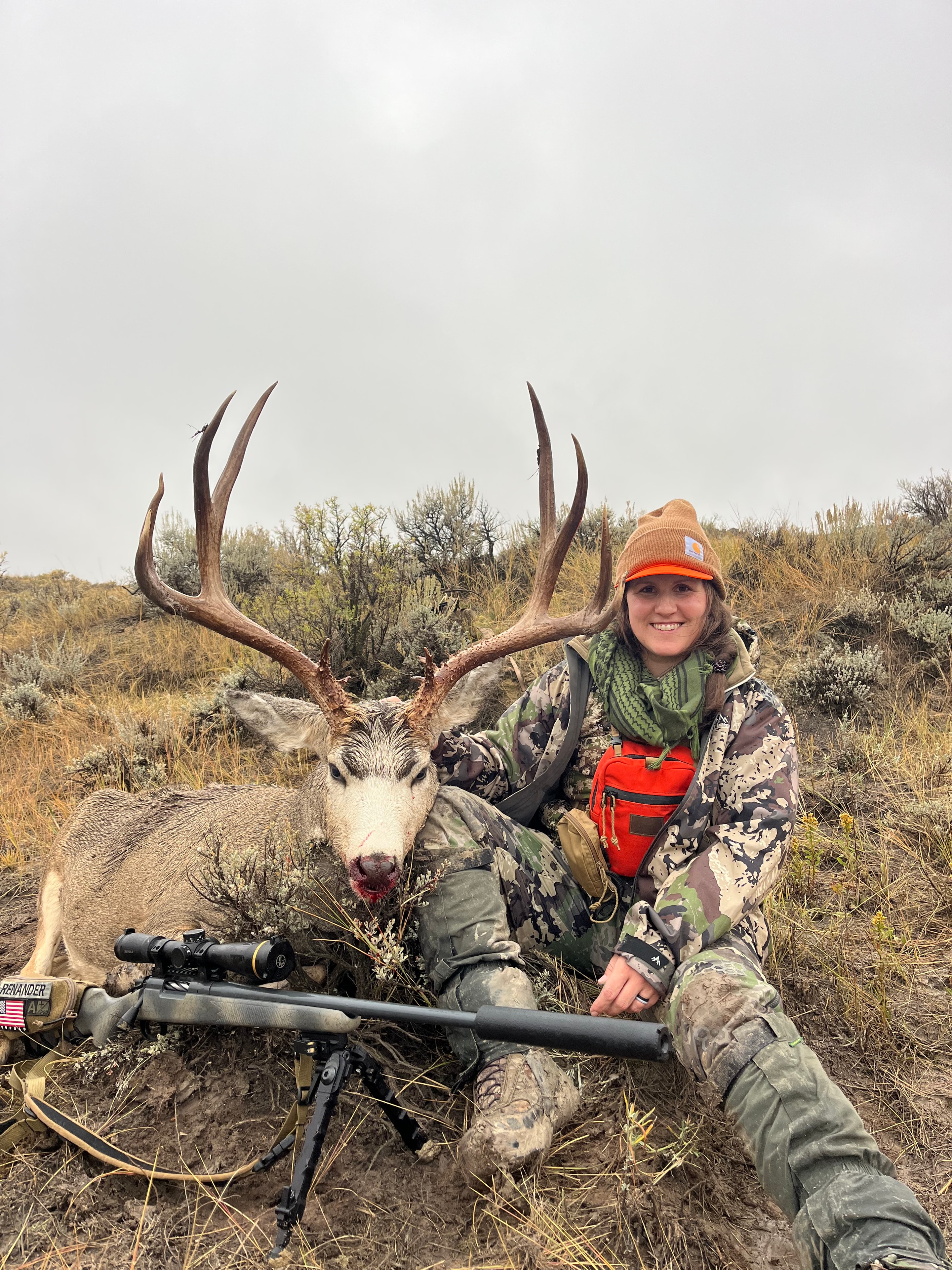 Two hunters with harvested mule deer buck and scoped rifle on bipod