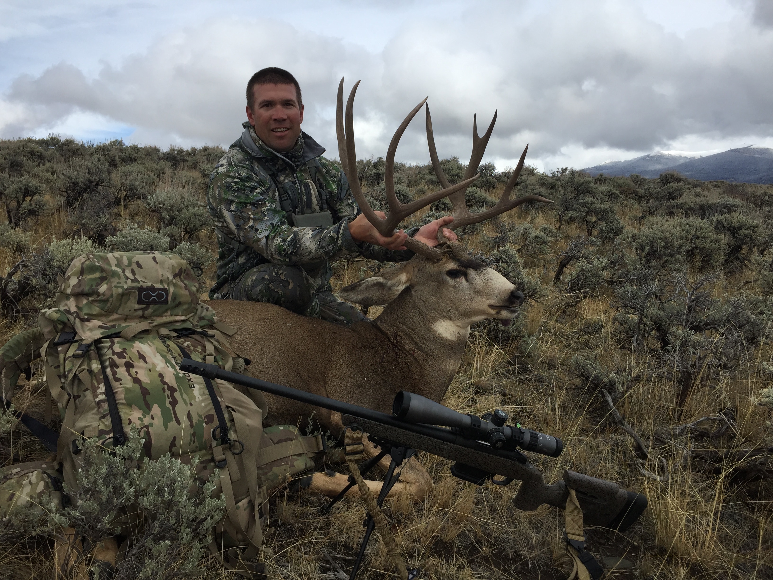 Hunter with mule deer buck and precision rifle in sagebrush, snowy peaks