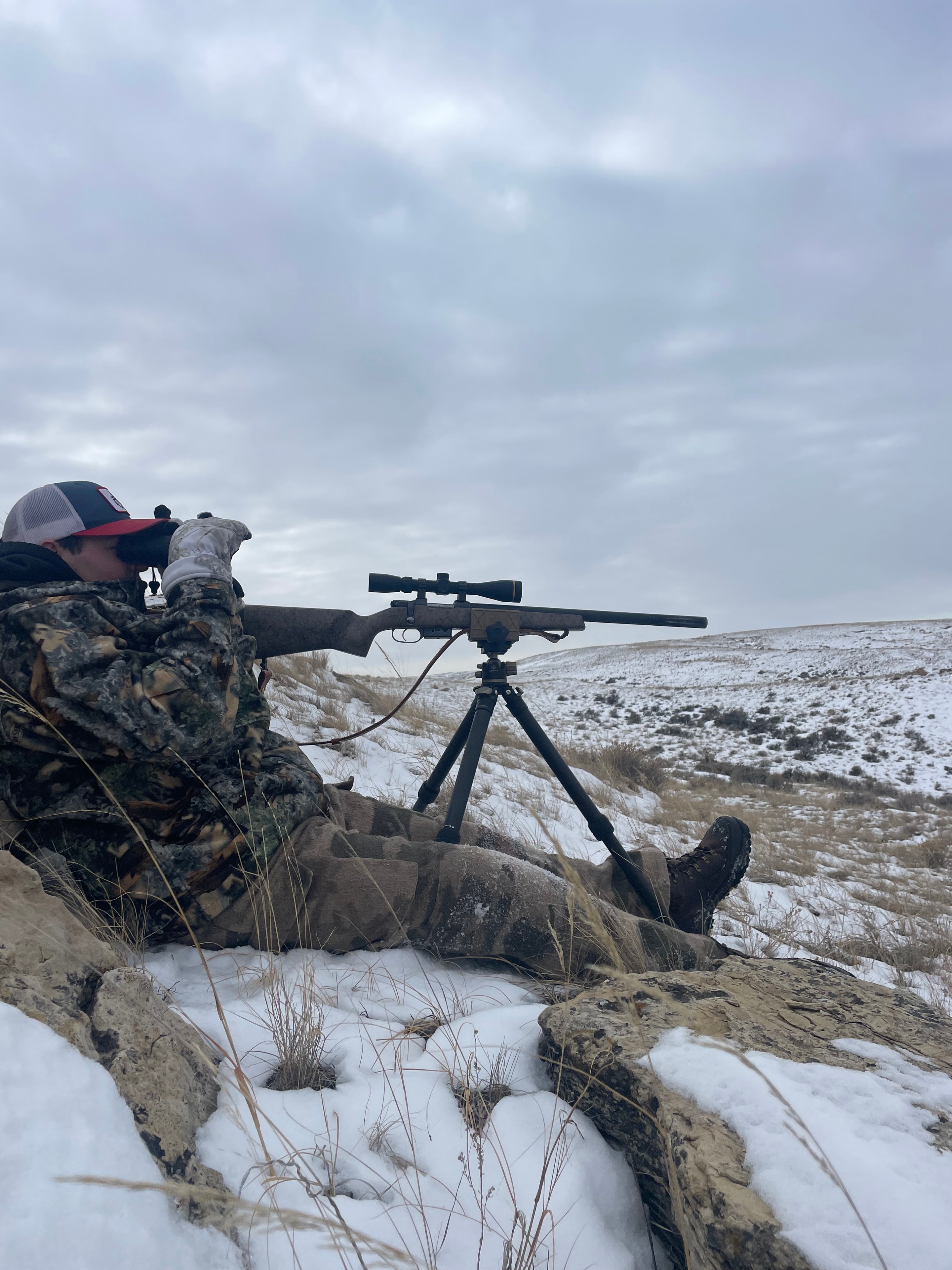 Hunter prone with scoped rifle on tripod in snowy badlands